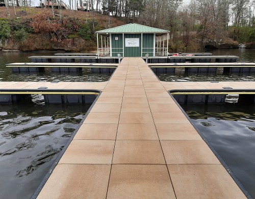 A wooden dock leads to a green-roofed building by a calm lake, surrounded by trees and empty boat slips.