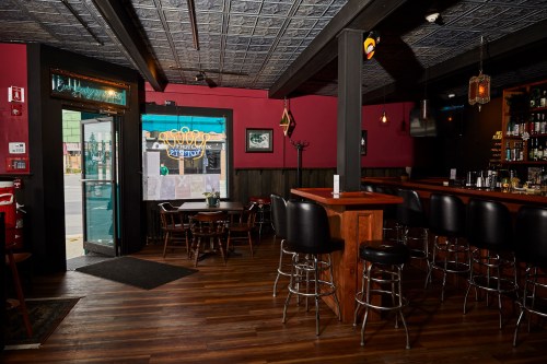 Cozy bar interior with wooden floors, black stools, and a view of the street through an open door.
