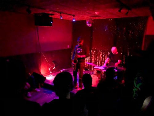 A dimly lit music venue with a saxophonist and drummer performing on stage, surrounded by an audience.
