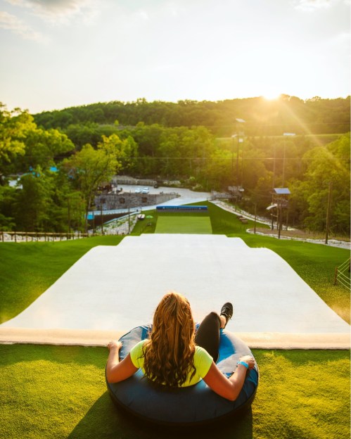 A person sits on a tube at the top of a grassy slope, ready to slide down as the sun sets in the background.