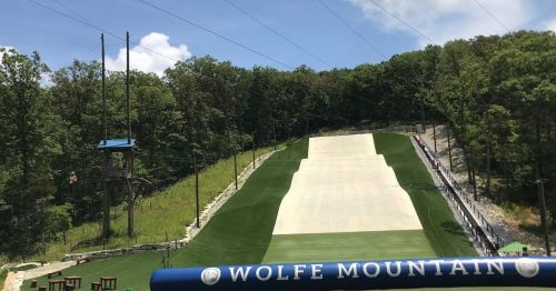 A view of Wolfe Mountain, featuring a grassy slope and zip line, surrounded by trees under a clear blue sky.