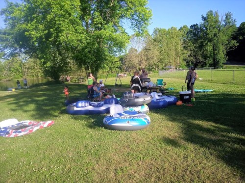 A group of people enjoying a sunny day by a river, surrounded by inflatable rafts and picnic items on the grass.