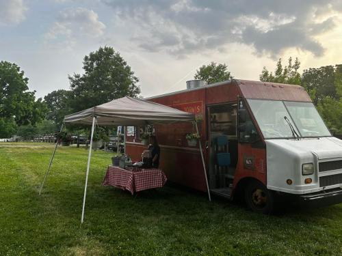 A food truck with a red exterior is parked on grass under a canopy, with a person preparing food outside.