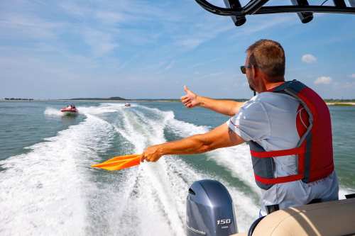 A man in a life jacket signals with an orange paddle while boating on a sunny day, with other boats in the background.