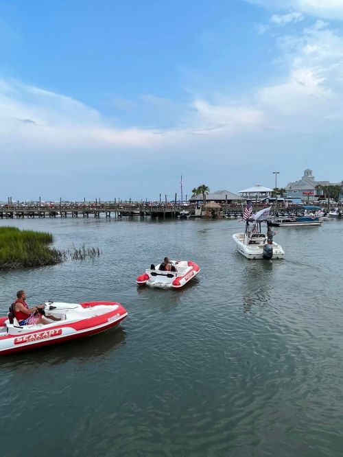 Two boats navigate a calm waterway near a marina, with a cloudy sky and greenery in the background.