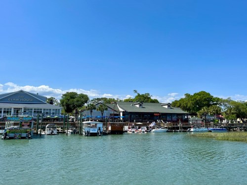 A waterfront view of a restaurant with boats docked, surrounded by trees and a clear blue sky.