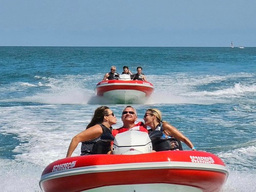 Two women kiss a man on a speedboat, while another boat with four people speeds behind them on the water.