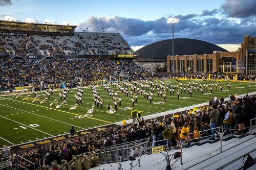 A large crowd watches a marching band perform on a football field at sunset, with stadium seating in the foreground.