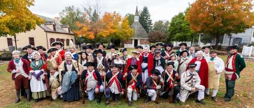 A large group of people in historical costumes pose outdoors, surrounded by trees with autumn foliage and historic buildings.