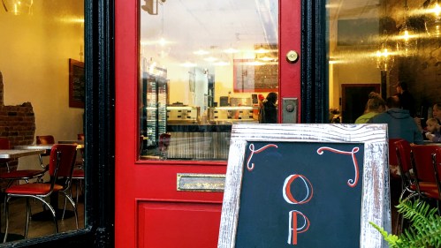 A red door leads into a cozy café with tables and a chalkboard sign outside.
