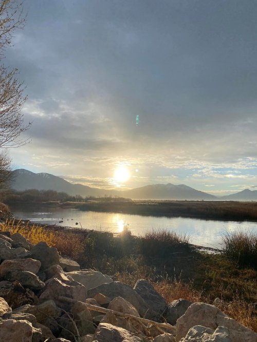 A serene landscape featuring a river reflecting the sun, surrounded by mountains and grassy banks under a cloudy sky.