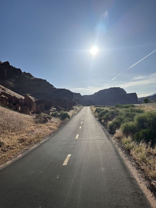 A sunlit road stretches into the distance, flanked by rocky hills and green grass under a clear blue sky.