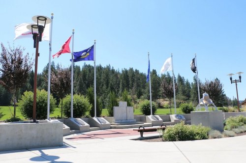 A memorial park with flags and greenery, featuring a stone monument and benches under a clear blue sky.
