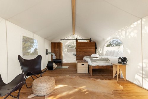 Cozy glamping tent interior with a bed, seating area, and wooden accents, surrounded by natural light.