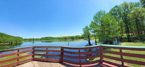 A scenic view of a calm lake surrounded by lush green trees under a clear blue sky, seen from a wooden deck.