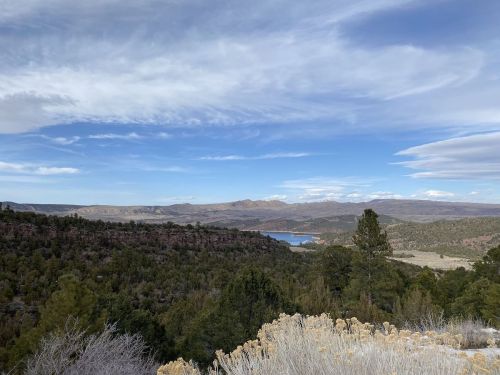 A scenic view of a mountainous landscape with a lake, surrounded by trees and a blue sky with clouds.