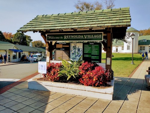 Welcome sign for Reynolda Village with a map and information board, surrounded by colorful plants and a sunny pathway.