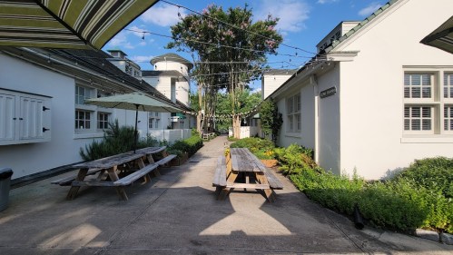A sunny courtyard with wooden picnic tables, greenery, and string lights between white buildings.