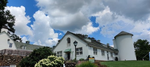 A white building with a round tower, surrounded by greenery and under a blue sky with fluffy clouds.