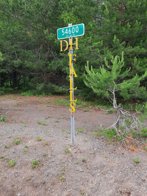 A green street sign with the number 54600 and the words "DH Falls" in yellow, surrounded by trees and gravel.