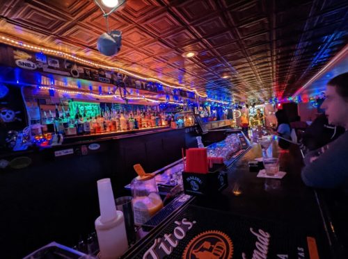 A vibrant bar scene with colorful lights, shelves of liquor, and patrons enjoying drinks at the counter.