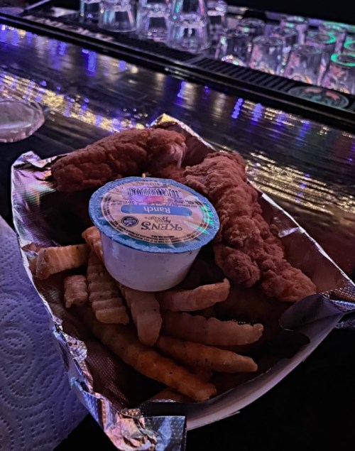 A plate of crispy chicken tenders with crinkle-cut fries and a side of ranch dressing on a bar counter.