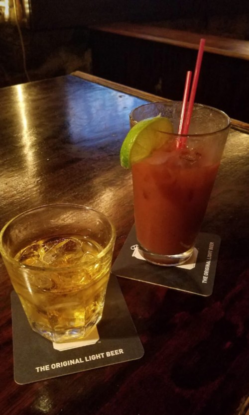 A glass of whiskey on the rocks and a Bloody Mary with a lime wedge, both on coasters on a wooden bar.