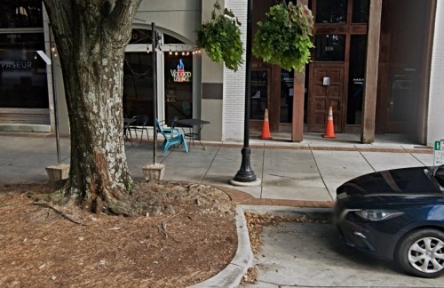 A tree stands near a sidewalk with outdoor seating, a restaurant entrance, and traffic cones in the background.