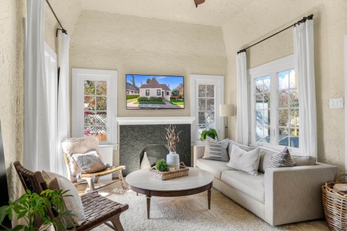 Bright living room with a cozy sofa, round coffee table, and a TV displaying a house exterior. Natural light floods in.