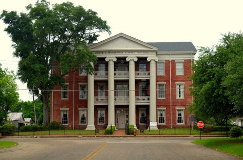Historic brick building with columns, surrounded by trees, featuring a stop sign in front and a grassy area.