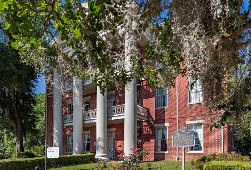 Historic brick museum building with white columns, surrounded by greenery and Spanish moss, under a clear blue sky.