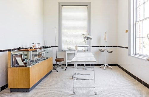 A bright, empty medical examination room with a glass display case, examination table, and medical equipment.