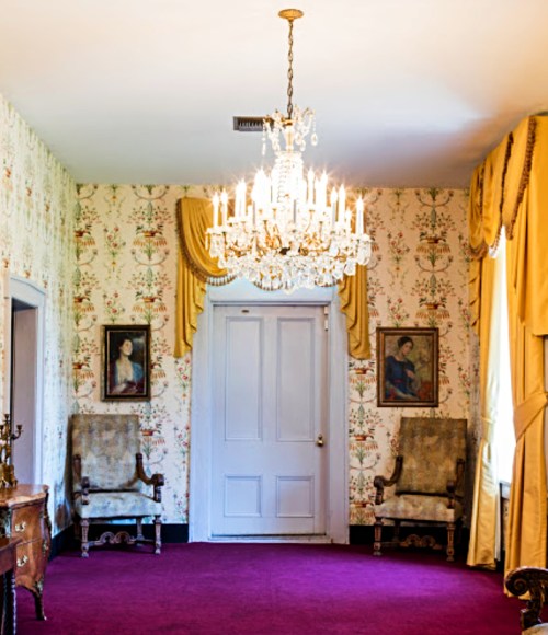 A vintage room with floral wallpaper, a chandelier, two armchairs, and paintings on the walls. Richly colored carpet.