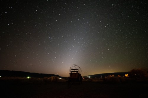 A silhouette of a wagon against a starry night sky, with faint lights in the distance and mountains in the background.