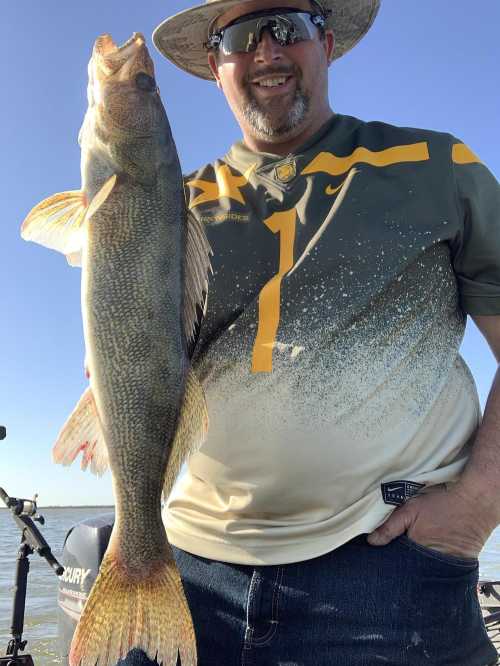 A smiling man in a hat holds a large fish, standing on a boat with a clear blue sky in the background.