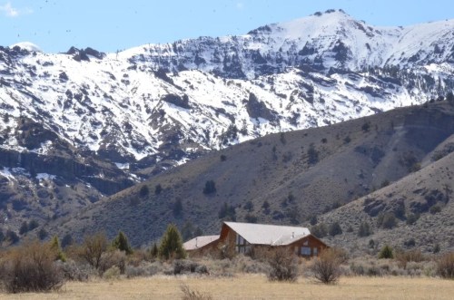 A wooden cabin nestled in a valley, surrounded by snow-capped mountains and sparse vegetation under a clear blue sky.