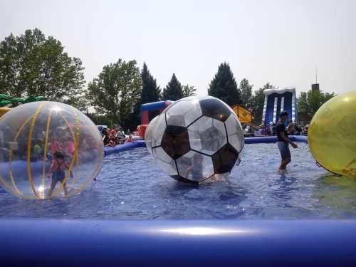 Children play in a water-filled inflatable area with large beach balls and a slide in the background on a sunny day.