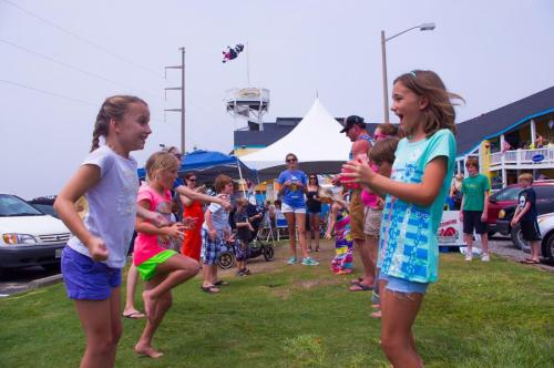 Children playing and dancing outdoors at a festive event, with colorful tents and a crowd in the background.