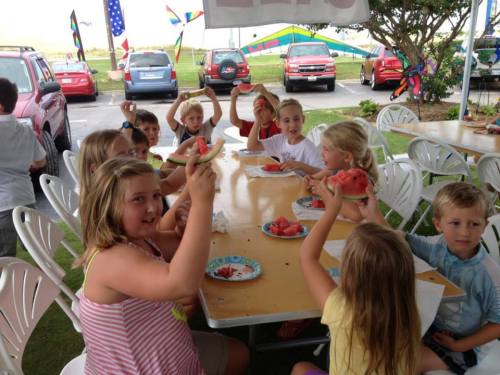 A group of children sitting at a table, joyfully holding up slices of watermelon during a summer gathering.