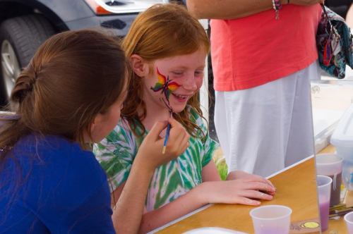 A girl with red hair smiles as another person paints a colorful design on her face at an outdoor event.