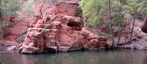 A large red rock formation beside a calm river, surrounded by lush green trees in a serene natural setting.