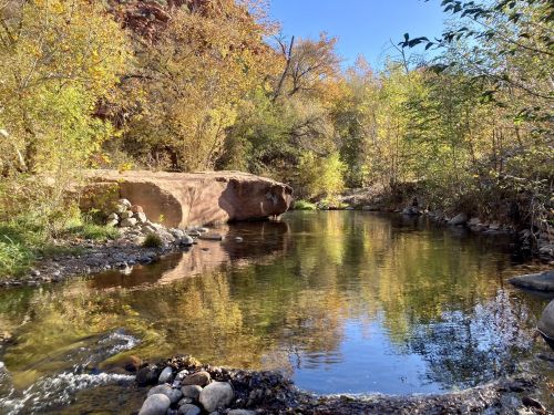 A serene river flows through a forest with autumn foliage, reflecting the colorful trees and clear blue sky.