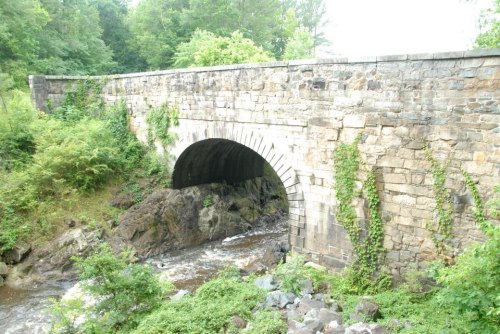 Stone bridge arching over a rocky stream, surrounded by lush greenery and trees.