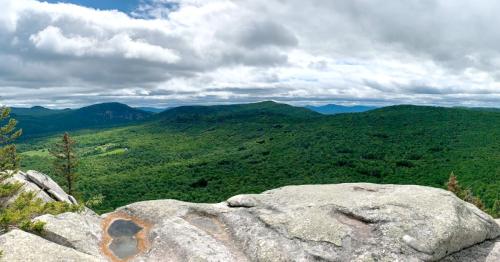 Panoramic view of lush green mountains under a cloudy sky, with rocky foreground and distant hills.