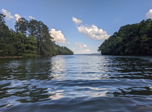 A serene lake scene with calm waters, surrounded by lush green trees and a blue sky with fluffy clouds.