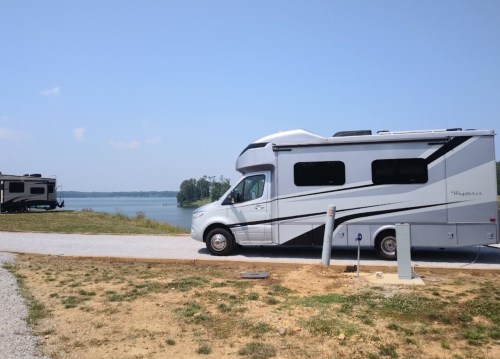 A silver RV parked by a lake, with another RV in the background and clear blue skies above.