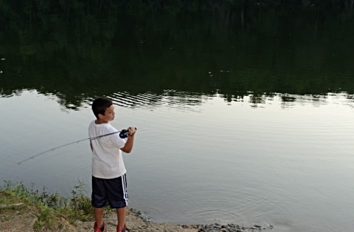 A young boy stands by a calm lake, casting a fishing line into the water. Trees reflect on the surface.