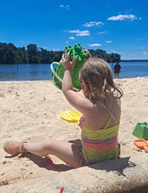 A young girl in a colorful swimsuit plays with a green sand bucket on a sandy beach by a lake on a sunny day.