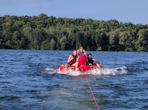 Four people enjoying a ride on a red inflatable tube being pulled on a lake, surrounded by trees and blue sky.