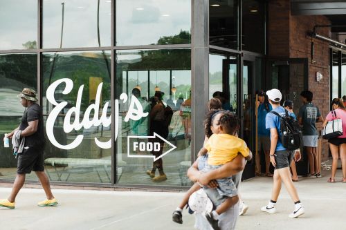 A busy entrance to Eddy's Food, with people entering and exiting, and a child being carried by an adult.